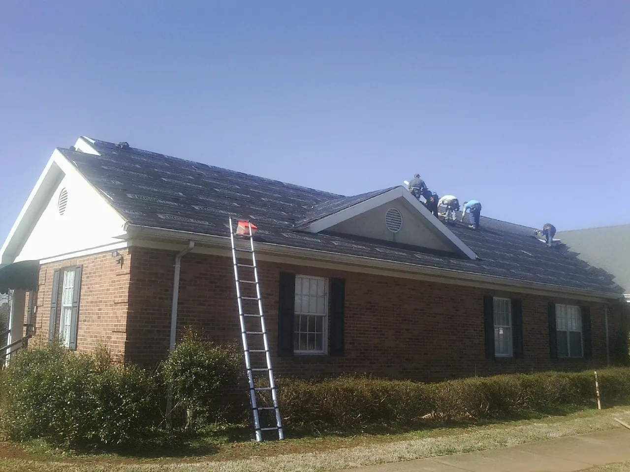 Crew tearing shingles off a brick ranch roof with a ladder set — Metal Roof Repair work by our crew in Amana