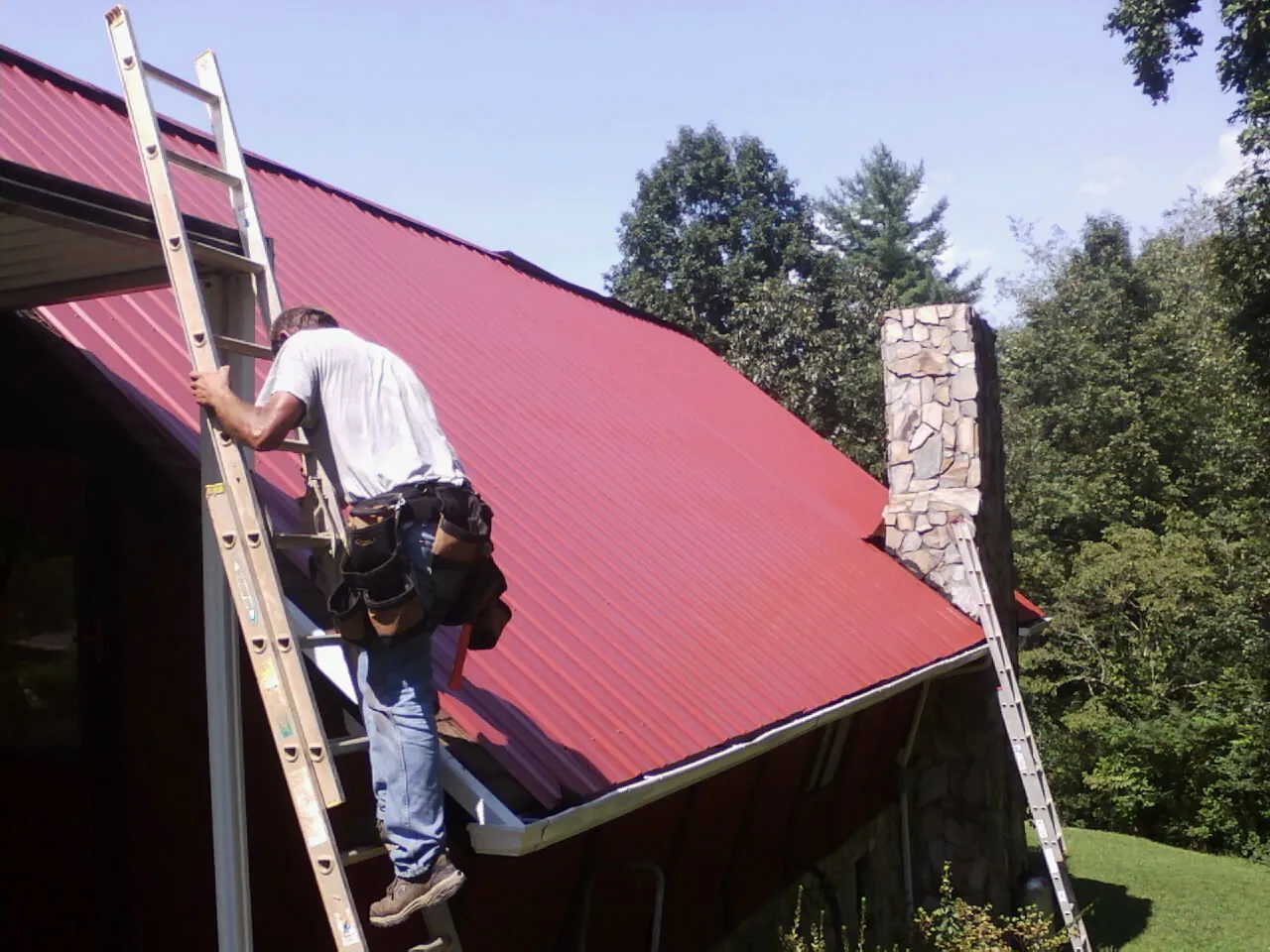 Roofer on a ladder beside a red metal roof and stone chimney — Soffit & Fascia Repair by Roofing Brothers in Amana